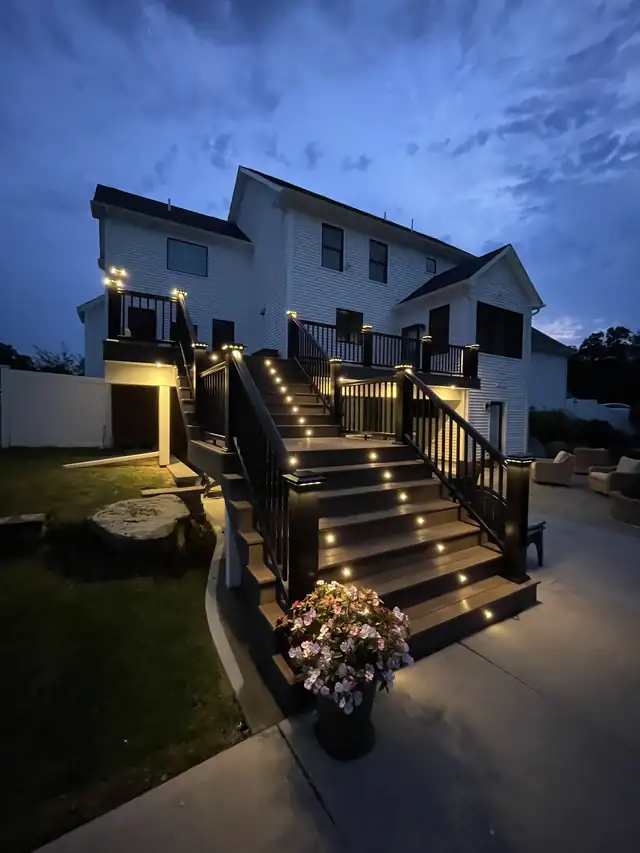 Lit composite deck stairs with black railings behind a white two-story home at dusk